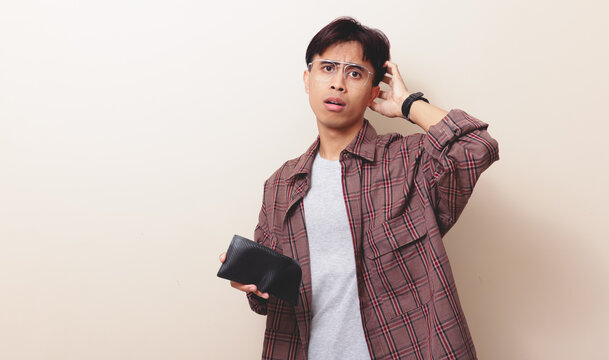 Confused young Asian man holding empty wallet with worried expression on isolated background.