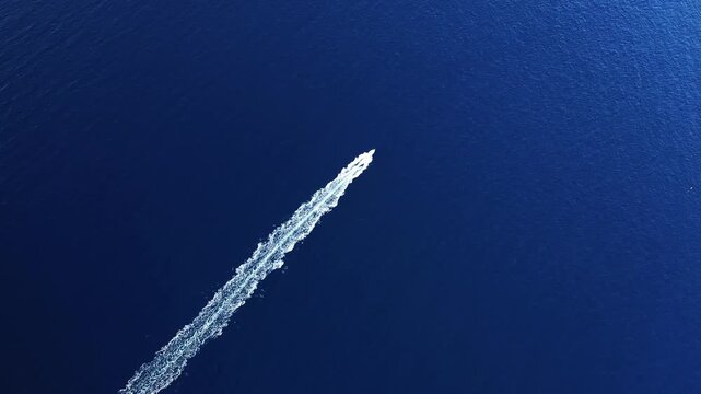 Aerial shot of a boat leaving a white trail on the deep blue sea near Calanques de Piana, Corsica. Minimalist composition emphasizing open water and movement.