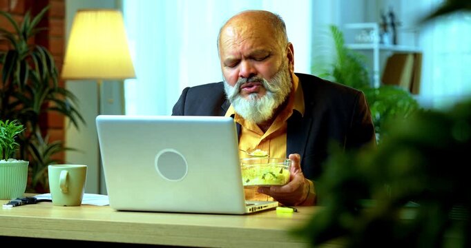 Indian senior businessman or corporate executive having a light snack while multitasking on a laptop and participating in a video call at his desk in a cozy office with moody lighting.