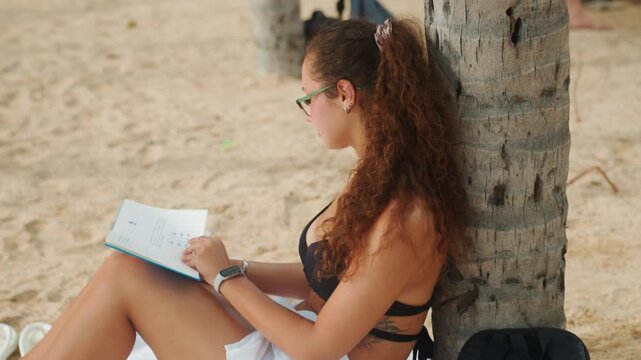 Woman studying notebook by palm tree seated on sand, focused notetaking while turning pages, gentle breeze and distant waves, backpack and pen at side, glasses and summer attire, blend