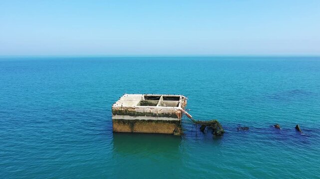 Aerial view of a concrete structure from the artificial harbor remains in the calm blue sea at Arromanches les Bains, Normandy, France. Clear sky and tranquil water create a peaceful coastal scene.