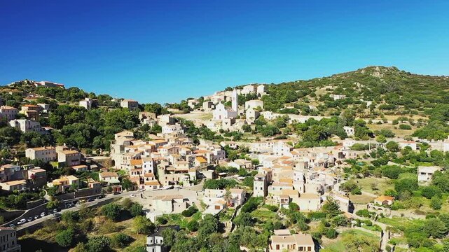Scenic aerial view of the hilltop village of Corbara surrounded by lush greenery and traditional Mediterranean architecture under a clear blue sky in Corsica, France.