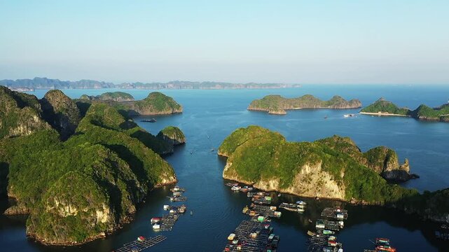 Aerial view of a floating village surrounded by lush green limestone islands in Ha Long Bay, Vietnam. Calm blue waters and scattered boats create a tranquil coastal landscape.