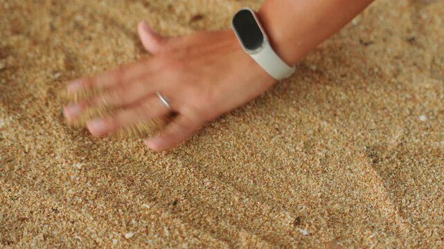 Hand tracing circles on sand beach, closeup of hand with watch and ring smoothing grains into concentric curves, gentle fingertip motion, warm sunlit texture and calming seaside atmosphere for mindful