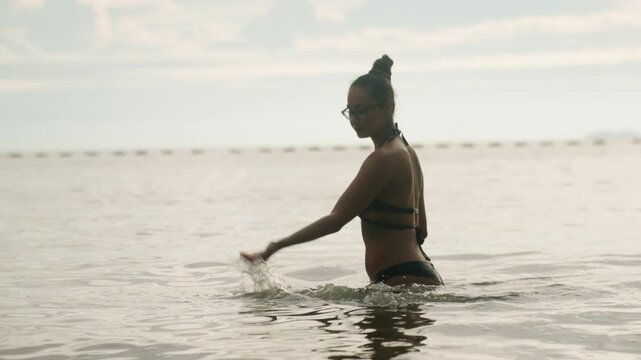 Woman wading in calm ocean, side profile with bun hairstyle and black swimsuit, soft sunrise light, gentle ripples around hips, distant horizon and muted sky creating contemplative seaside mood.