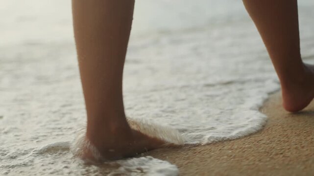 Bare feet walking on sandy shore, slow closeups of toes touching foamy waves, gentle step and lift, pausing to leave soft footprints in wet sand, warm golden light and coastal breeze evoke calm