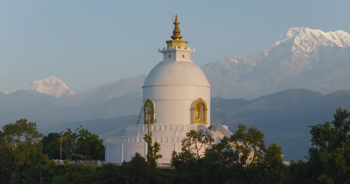 Aerial drone shot flying around the white World Peace Pagoda in Pokhara Nepal revealing a majestic backdrop of the snow-capped Annapurna and Machhapuchhre mountain peaks in the Himalayas.