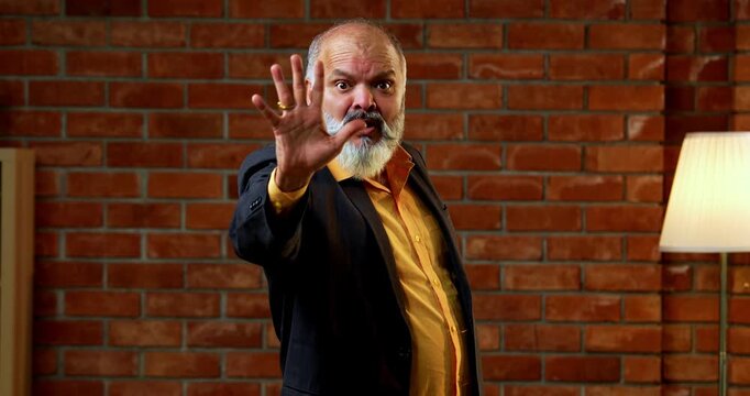Agitated Indian senior businessman standing against a red brick wall in a cozy office, gesturing a stop sign with his hand and showing a frustrated or serious expression during a conflict.