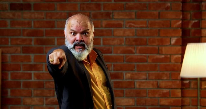 Agitated Indian senior businessman standing against a red brick wall in a cozy office, gesturing a stop sign with his hand and showing a frustrated or serious expression during a conflict.