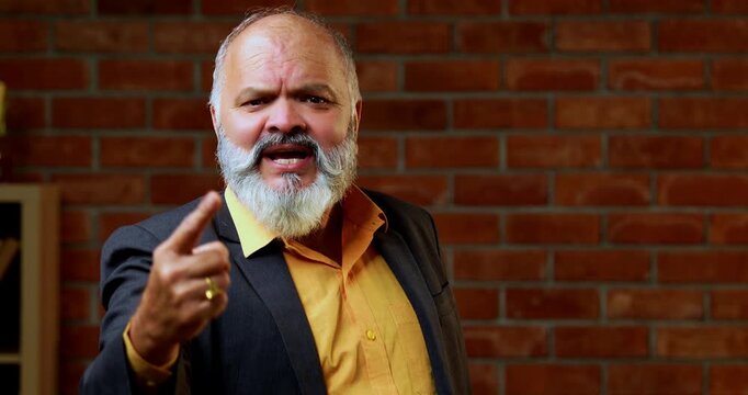 Angry Indian senior businessman standing against a red brick wall in a cozy office, scolding and looking directly at the camera with a fierce, frustrated expression to show authority or conflict