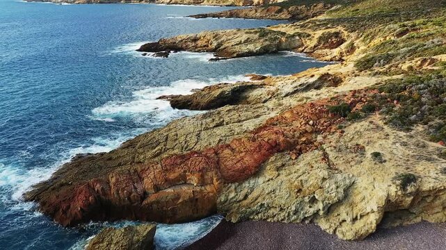 Dramatic rocky shoreline with waves crashing against the coast at Delta du Fango, Corsica. Rugged natural landscape with blue sea and textured cliffs.