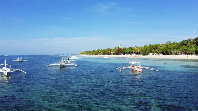 Traditional outrigger boats float on clear blue water near a white sandy beach lined with palm trees on Panglao Island, Philippines.