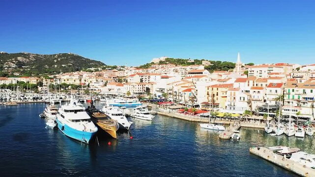 Scenic view of luxury yachts docked in the vibrant marina of Calvi, Corsica, with colorful Mediterranean buildings and hills in the background under a clear blue sky.