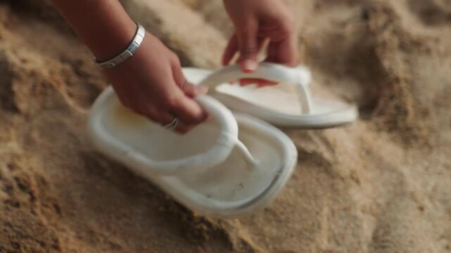 White sandals lying on sandy beach, person retrieves pair with tender hands, closeup textured sand and fading footprints, warm golden light, calm nostalgic mood and quiet retreat by shore
