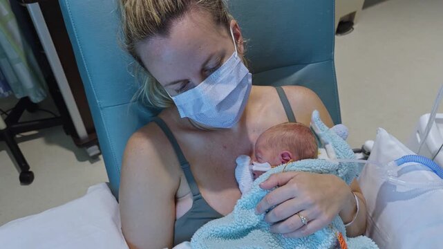 Young mother wearing surgical mask holds tiny premature newborn wrapped in blue blanket in hospital recliner chair in NICU.