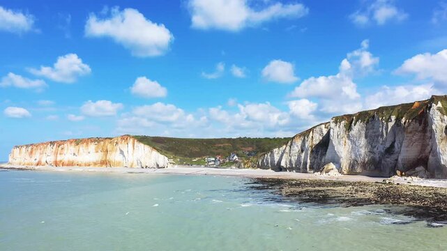 Scenic view of dramatic white chalk cliffs and beach at Les Petites Dalles, Normandy, France, under a bright blue sky with scattered clouds.