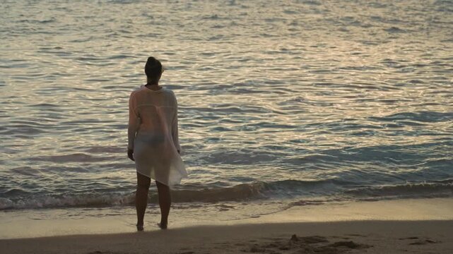 Woman faces gentle morning sea, soft dawn light illuminates rippling water, light dress brushing knees, subtle movement as waves touch bare feet, calm steady gaze suggesting renewal and resolve,