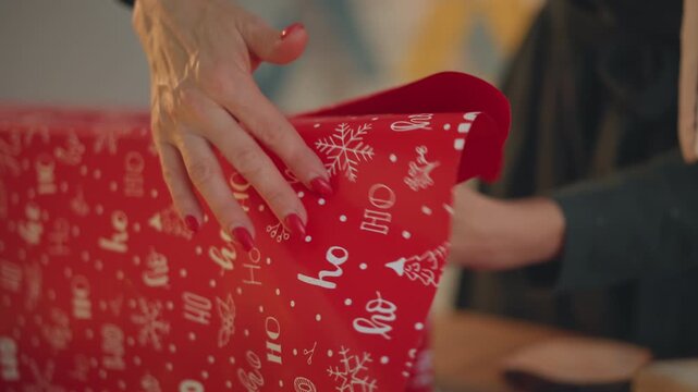 Experienced hands wrapping christmas present on table, large box wrapped with patterned red paper, quick precise folds, tucking and sealing, skillful movements, instructional home scene, preparation