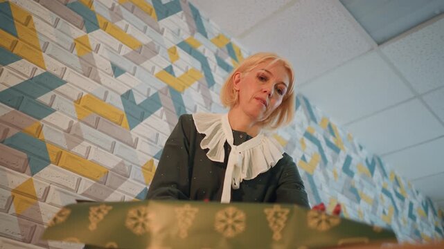 White woman wrapping christmas present in boutique, retro blouse with ruffle collar, patterned wallpaper backdrop, lowangle overhead shot, green gift box with gold motifs, careful hands tying ribbon,