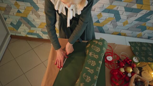 Caucasian woman wrapping christmas present on kitchen table surrounded by ribbons, ornaments, scissors, tape and fairy lights, hands cutting paper and tying bow, warm cozy holiday mood and handmade