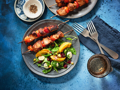 The image shows grilled meat skewers alongside a fresh salad with peach and nuts on a blue table. This scene is perfect for restaurant menus, food blogs, or social media posts promoting summer dining