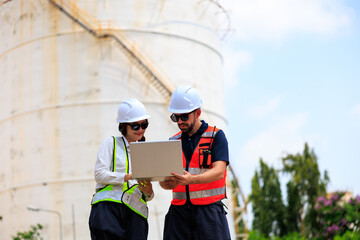 A diverse team of male and female industrial engineers in safety gear discussing project data on a laptop outside a petrochemical plant with large white storage tanks. © Nalinee