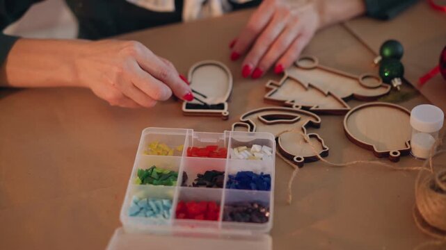 Closeup hands crafting wooden christmas ornaments on table with beads, ribbon, glue female with red manicure placing colorful beads into ornament frames warm festive lighting, cozy holiday DIY mood,
