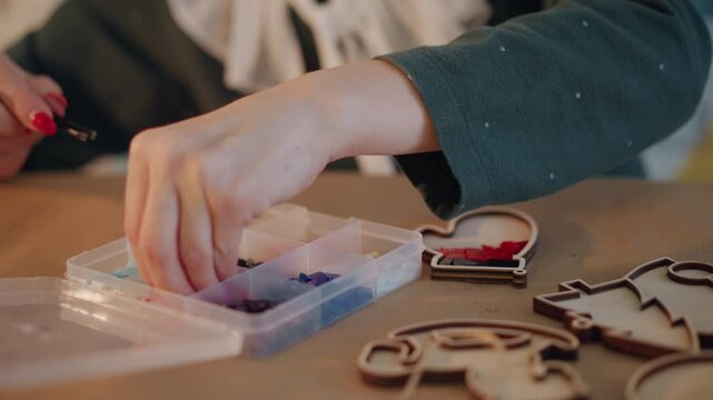 Closeup hands sorting colorful craft beads on wooden table, organizer box open, wooden ornament shapes scattered, tweezers picking tiny beads, warm cozy lighting, tranquil holiday workshop preparing