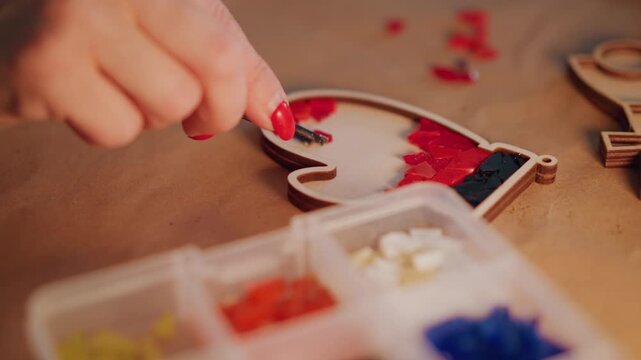 Closeup hands filling heart ornament with red beads, mosaic pattern forming inside wooden frame, tweezers placing tiny pieces with steady motion, warm glow and intimate festive atmosphere, final