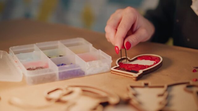 Closeup hands crafting christmas heart present, artisan picking tiny beads with tweezers on wooden table, red nails, warm lamp glow, DIY gift tag scene, detailed texture and soft bokeh