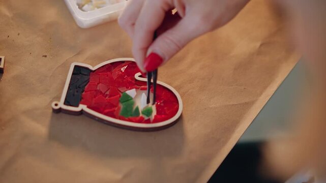 Closeup christmas mitten ornament being assembled by female hand with tweezers, red nail polish placing green tile into red mosaic, cozy tabletop craft workshop, holiday gift making atmosphere