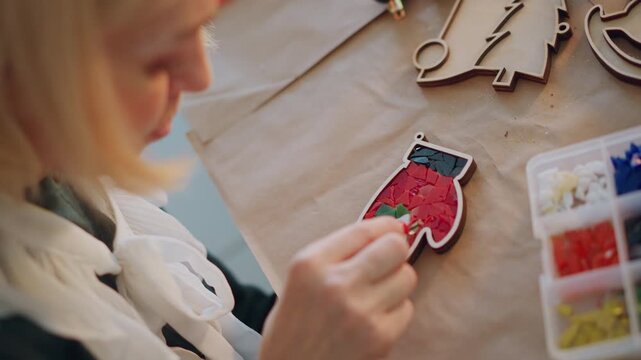 White woman crafting christmas present embroidering mitten ornament with colorful beads, closeup hands arranging bead tray on wood table, warm natural light, cozy holiday workshop vibe, scarf wrapped