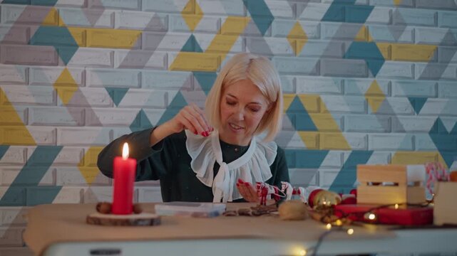 Caucasian woman wrapping christmas present by candlelight on cozy table with ribbon scissors small boxes fairy lights patterned wall focused senior smiling crafting festive decoration warm glow
