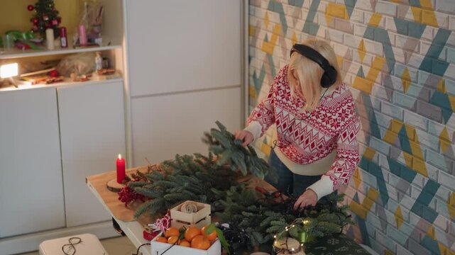 White woman wrapping Christmas presents at table upbeat playlist, headphones, patterned sweater, string lights, scissors, ribbon and tags, busy gift station in modern kitchen, fast festive prep