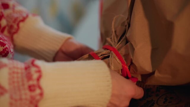 Caucasian hands tie ribbon on christmas present, focused closeup of knot being formed with patterned sweater sleeves, brown kraft paper and warm bokeh lights, finishing touch before gift exchange