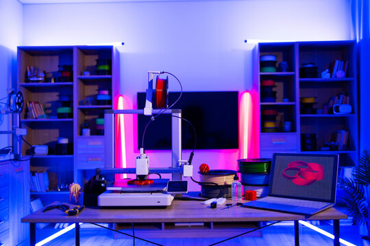 A modern 3D printer is set up on a desk with spools of filament and a laptop displaying a 3D model, illuminated by vibrant neon lighting
