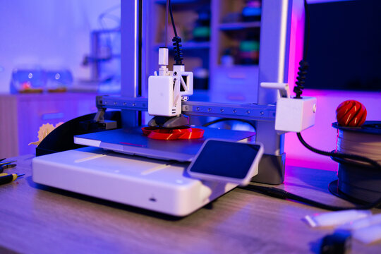 A modern 3D printer is actively creating a red plastic object on its build plate with filament spool nearby