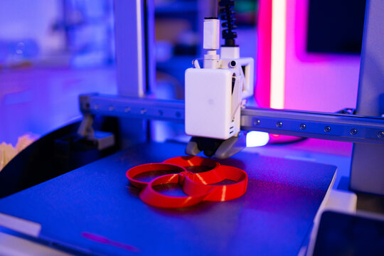 A modern 3D printer is actively creating a red object layer by layer with vibrant blue and pink lighting