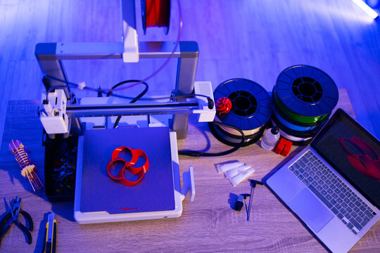 A 3D printer is actively creating a red object on its build plate, surrounded by spools of filament, tools, and a laptop displaying a 3D model