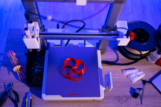 A 3D printer is actively creating a red, intricate object on its build plate, surrounded by filament spools and tools