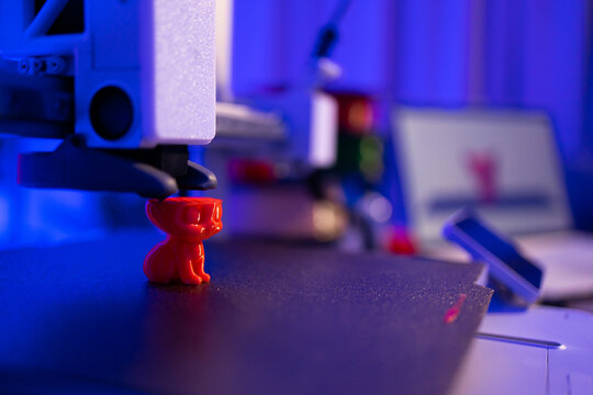A 3D printer nozzle extrudes molten plastic to create a small red cat figurine on its build plate with a laptop in the background