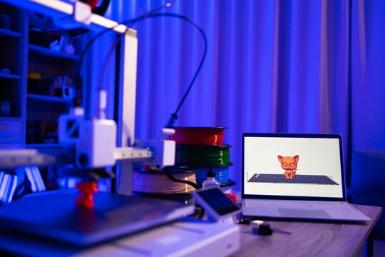 A 3D printer is actively creating a small orange cat figure, with spools of filament and a laptop displaying the 3D model nearby
