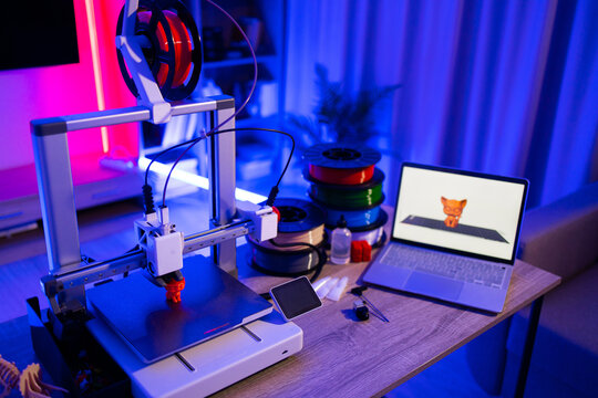 A 3D printer is actively creating a small orange object while a laptop displays its digital model, surrounded by spools of filament and tools