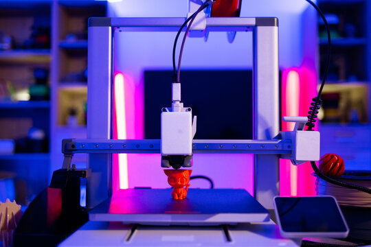 A modern 3D printer is actively creating a small orange cat figurine on its build plate, illuminated by vibrant neon lights