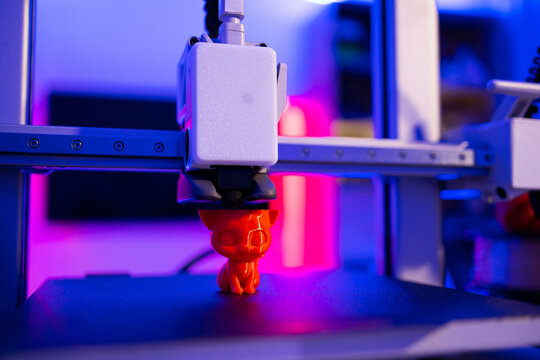 A 3D printer is actively creating a small, orange cat figurine with intricate details, illuminated by vibrant blue and pink lights