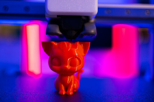 A red 3D printer nozzle extrudes filament to create a small, shiny red cat figurine with vibrant pink and blue lighting