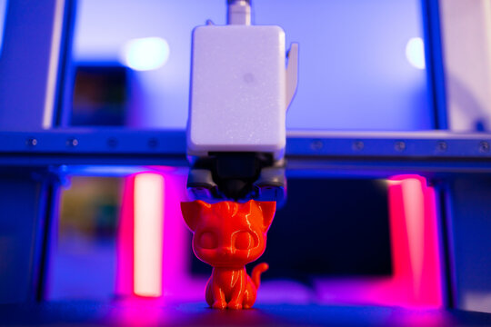 A 3D printer is actively creating a small, bright red cat figurine with intricate details under vibrant blue and pink lighting