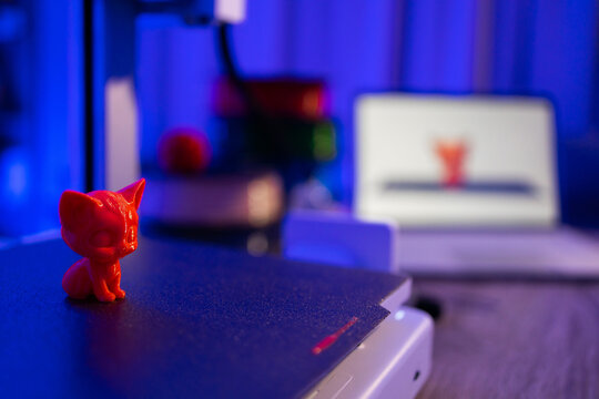 A small red 3D printed cat figurine sits on a 3D printer bed with a laptop displaying the printer software in the background