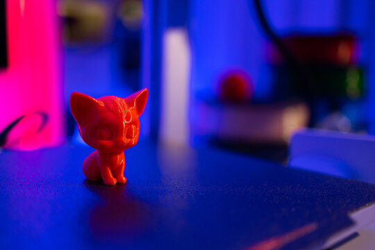 A small, bright red cat figurine sits on a dark surface illuminated by vibrant neon pink and blue lights, creating a futuristic atmosphere