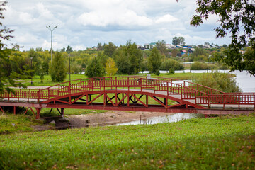 Fototapeta premium Red wooden bridge over a small river surrounded by lush greenery and trees under a cloudy sky in a tranquil park setting